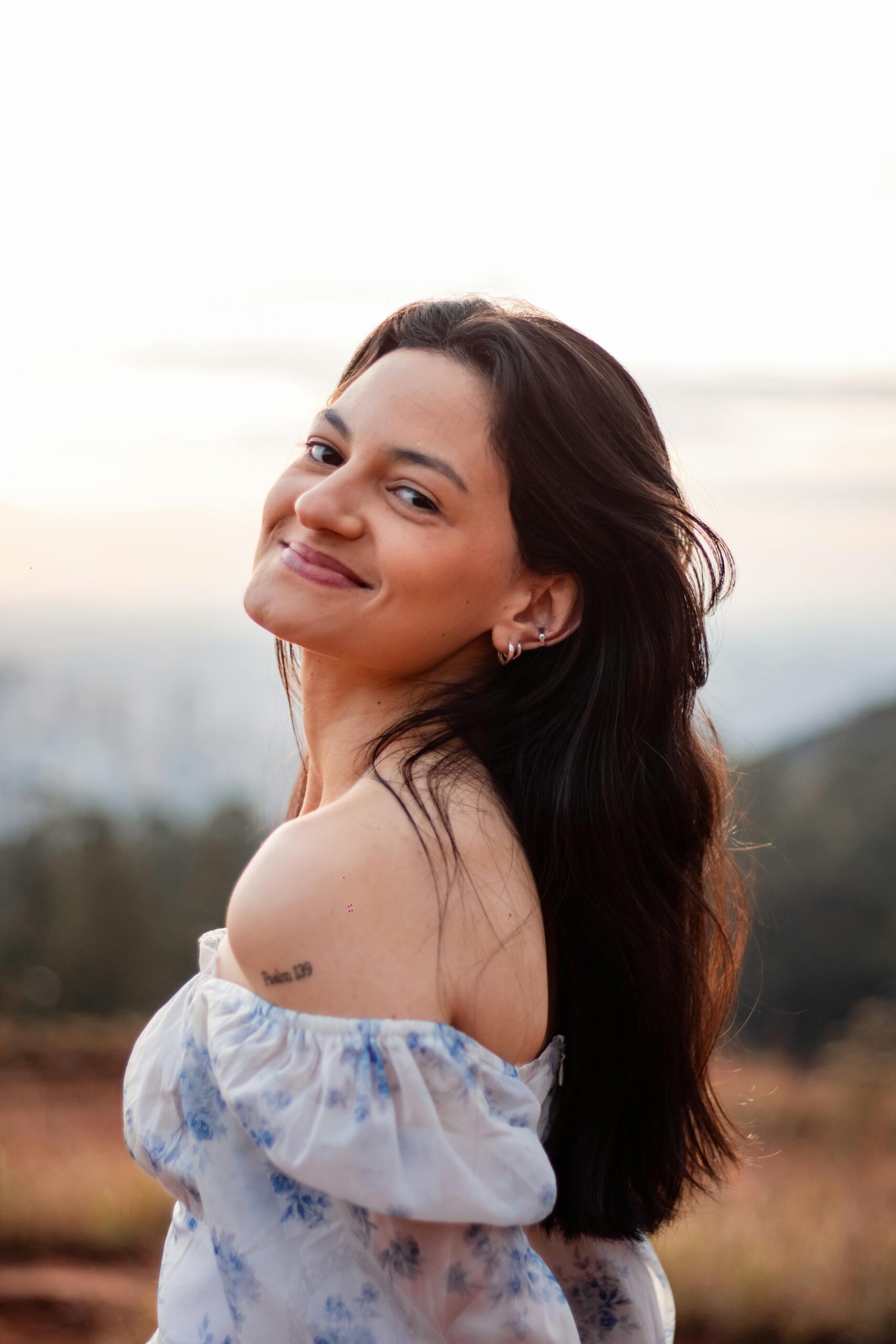 A young woman smiles warmly in an outdoor setting wearing a floral off-shoulder top.